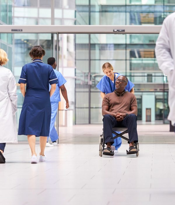 Female Nurse Wearing Scrubs Wheeling Patient In Wheelchair Through Lobby Of Modern Hospital Building